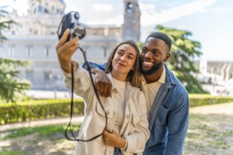 Happy multi-ethnic couple taking selfie with digital camera in a park with views on downtown