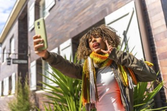 Low angle view of a cheerful latin young woman with curly hair taking selfie outside a residential
