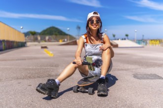 Young female asian influencer sitting on skateboard park holding mobile phone under the sun