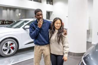 Happy multiethnic couple choosing their new electric car in a dealership showroom, sustainable