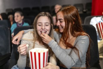 Two female friends enjoying a movie night, sharing popcorn and laughter in a crowded cinema