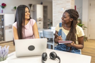 Two smiling teenage girls collaborating on school project, using laptop and taking notes, enjoying