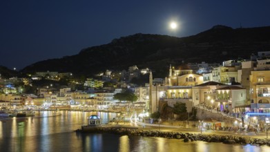 Illuminated coastal town at night with moon in the sky and water in the foreground, Rising full
