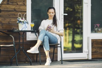Relaxed woman sitting on terrace of wooden a-frame cabin with smartphone