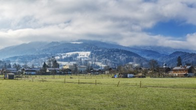 A village in front of snow-covered mountains under a cloudy sky on a green meadow, Bad Feilnbach,