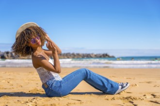Beauty latin woman sitting on the sand of a beach playing with sun hat and smiling at camera