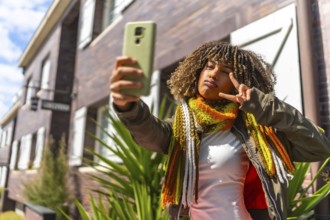 Beauty latin woman gesturing victory while taking a selfie standing next to a house