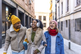 Three cheerful multi ethnic young friends walking arm in arm in the city on a winter day, enjoying