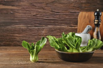 A bunch of vibrant bok choy sits in a bowl alongside a single piece on a wooden table, complemented