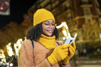 Smiling young woman wearing yellow winter clothes using smartphone in a city street at night with