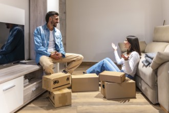 Young couple talking and smiling while unpacking cardboard boxes in new home, planning the
