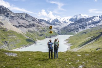 Two friends throwing yellow hats in the air, mountaineers in front of a blue mountain lake, Lac des