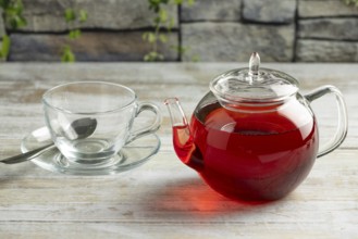 A teapot with red tea next to an empty glass cup with spoon on a table in front of a stone