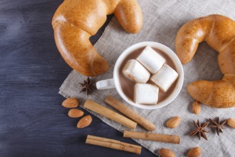 A cup of hot chocolate with marshmallow, croissants and spices on linen napkin and black wooden