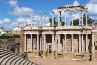 Ancient Roman theatre with columns and stairs, in the background a modern city under a blue sky,