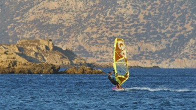 Person windsurfing in front of rocky coastal scenery and blue mountains in the background,