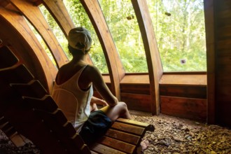 A woman is sitting on a wooden bench in a forest. She is wearing a white tank top and blue shorts.