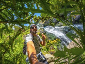 A man is taking a picture of himself in a forest. He is wearing a hat and a backpack