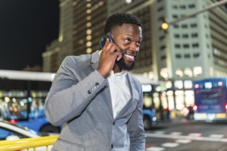 Young businessman smiling while talking on his smartphone in a bustling city street at night,