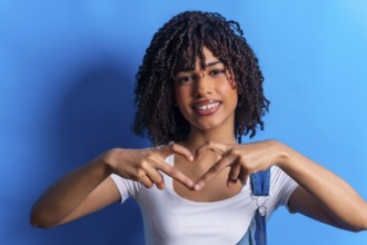 Smiling young woman making heart shape with hands, expressing love and affection on vibrant blue