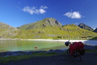 View of a bicycle with red bags on the shore of a bay, surrounded by green hills and mountains