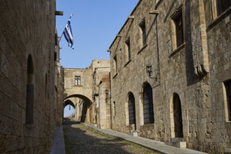 Historic street with stone buildings and arches, decorated with a Greek flag on a sunny day,