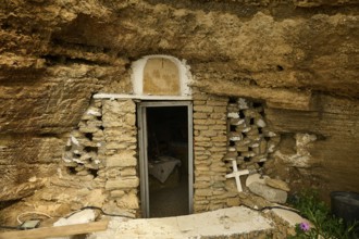 Entrance to a cave carved into the rock, surrounded by a stone wall, with a cross and plants on the