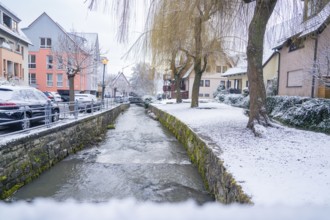 A small river flows through a snow-covered village with houses, Aidlingen, Böblingen district,
