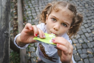 A young girl with pigtails eats a snack on a cobbled street and looks curiously, Stuttgart,