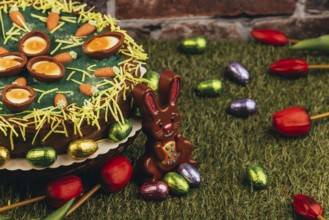 An Easter cake with carrots next to chocolate eggs, Easter bunnies and tulips on green grass