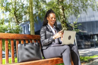 Latin elegant businesswoman using mobile phone while working with laptop on remote sitting outdoors