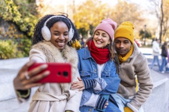 Three happy multi ethnic friends wearing winter clothes taking a selfie with a smartphone in a city