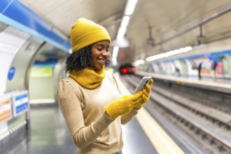 Cheerful young black woman wearing yellow winter clothes and using smartphone while waiting for