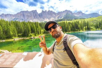 Tourist gesturing peace sign while taking a selfie at lake carezza with latemar mountain range in