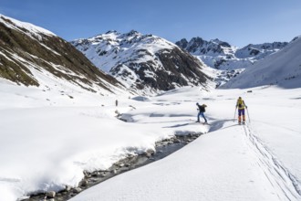 Two ski tourers in the snow crossing a mountain stream in the Funtauna Valley, Graubünden Haute