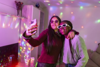 Two teenage girls wearing sunglasses are taking a selfie with a smartphone at a home party with