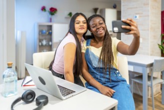 Two happy multi ethnic female students taking a selfie with a smartphone while studying at home
