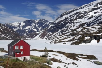 A red house stands isolated in a snow-covered mountain landscape under a clear sky, spring in the