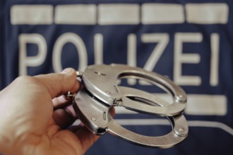 A pair of handcuffs is held by one hand, a police sign in the background