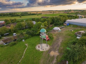 A tower stands in a rural area, surrounded by meadows, sheds and trees under a cloudy sky at