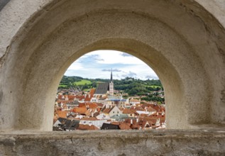 View of Ceský Krumlov town through a stone arch, featuring rooftops and church steeple, Cesky