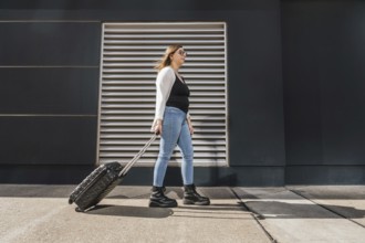 Young business woman pulling rolling luggage along a sunny city sidewalk, walking past a sleek