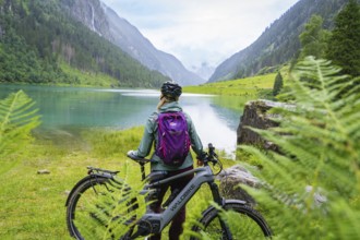 A woman with a helmet and purple rucksack stands next to an e-bike by the lake and looks out over
