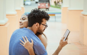 Unfaithful man looking at the cell phone while hugging his girlfriend outdoors. Unfaithful