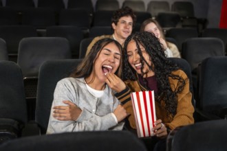 Two young women are enjoying a movie at the cinema, laughing and sharing popcorn, with other people