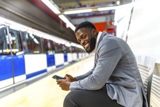 Young black businessman sitting on a bench, using a smartphone while waiting for a subway train at