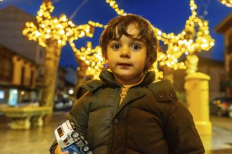A young boy is standing in front of a Christmas tree with a toy car in his hand. The scene is