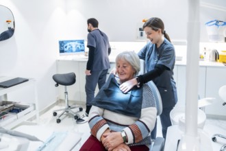 Dentist adjusting bib on senior woman for a dental checkup in a modern clinic, while a technician