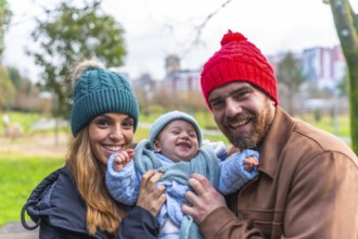 Smiling parents holding their laughing baby wearing winter clothes, enjoying a sunny winter day at