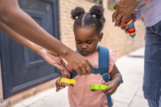 Baby african girl with schoolbag holding car toys standing in the street